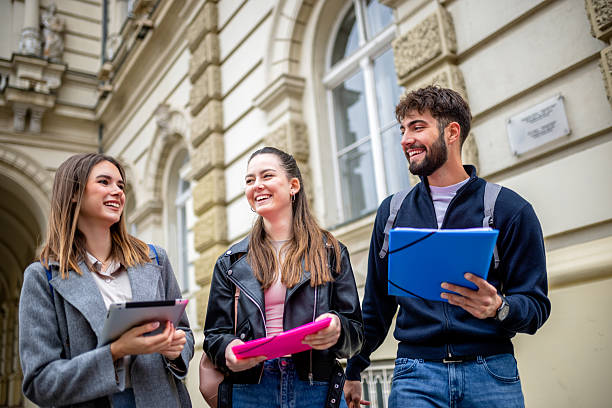 hree students stand together outdoors, holding books and literature, engaging in a lively conversation filled with laughter. Dressed casually and carrying backpacks.